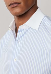 Uomo che indossa una camicia a righe verticali azzurra e bianca, con colletto bianco a contrasto, abbottonata al collo e al petto.