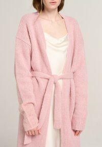 Helles rosa gestricktes Cardigan mit einem Taillengürtel, übergroßen Ärmeln und gerippten Bündchen, getragen über einem cremefarbenen Satin-Kleid. Weicher Stoff.