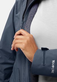 Donkerblauwe waterdichte jas met een voorkantsrits, fleece-voering bij de kraag en klittenbandsluitingen aan de mouwen. De stof ziet er soepel uit met een zachte textuur.