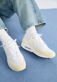 Vita Nike Air Max 1-sneakers med gula accenter, som har en blandning av släta och texturerade material, snörning och synlig luftkuddar.