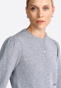 Femme aux yeux bleus portant des boucles d'oreilles en argent et un pull en tricot gris clair à boutons, sur un fond blanc uni.