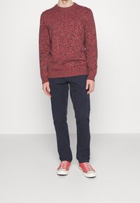 Maglione a maglia rosso mélange con scollatura rotonda, abbinato a pantaloni blu scuro e sneakers rosse. Presenta polsini e orlo a coste.