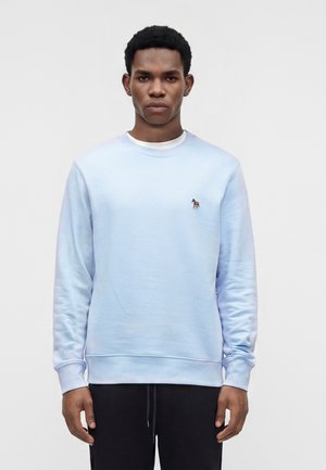 Jeune homme aux cheveux courts portant un sweat-shirt bleu clair avec un petit logo zèbre sur la poitrine et un pantalon noir, debout en position neutre.