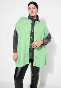 Mintgrön, oversized stickad kofta med silverknappar, tillsammans med en mörk denimskjorta. Modellen bär svarta läderliknande byxor.