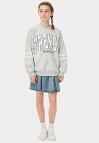 Felpa grigia con stampa "FRENCH RIVIERA" e dettagli floreali, abbinata a una gonna di jeans. Sneakers bianche e calzini leggeri completano il look.