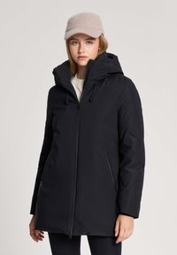 Młoda kobieta w czarnej damskiej kurtce z kapturem oraz beżowej wełnianej czapce, stojąca na tle jasnoszarego, gładkiego tła.