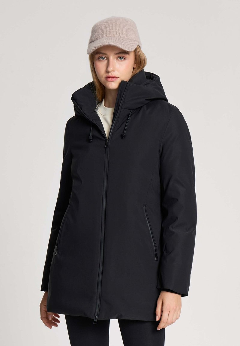 Młoda kobieta w czarnej damskiej kurtce z kapturem oraz beżowej wełnianej czapce, stojąca na tle jasnoszarego, gładkiego tła.