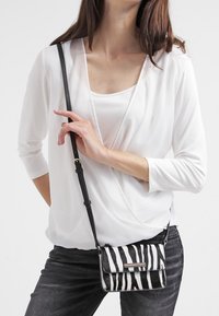 Sac bandoulière à imprimé zèbre noir et blanc avec une longue sangle, featuring un rabat avant et des accessoires en métal, porté sur un haut blanc.