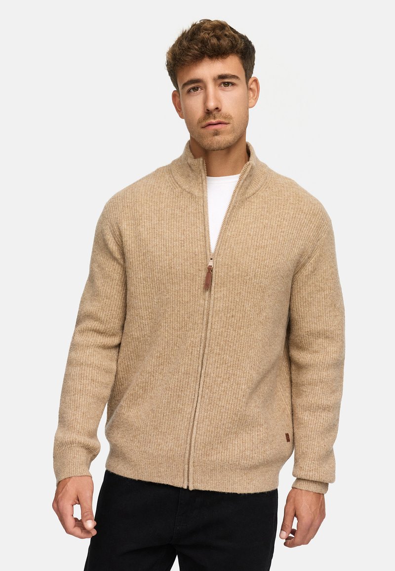 Beżowy sweter na zamek z materiału ribbed, z wysokim kołnierzem i brązowym skórzanym uchwytem zamka. Model nosi go na białej koszulce.