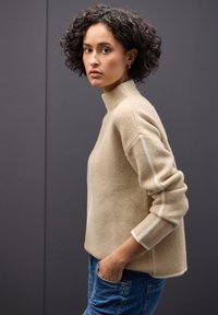 Beiger Strickpullover mit hohem Kragen, strukturiertem Stoff und weißen Akzentstreifen an den Ärmeln; getragen mit blauen Jeans.