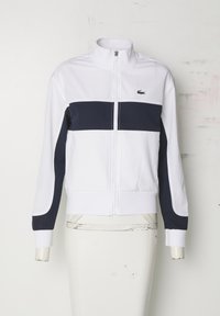 Chaqueta blanca con cierre y una franja horizontal azul marino en el pecho. Presenta un cuello alzado y puños elásticos. Logo de Lacoste en el pecho.