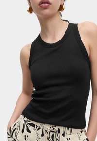 Zwarte geribbelde tanktop met een getailleerd ontwerp en mouwloze snit, gecombineerd met lichtgekleurde gebloemde broeken. Gouden geometrische oorbellen zichtbaar.