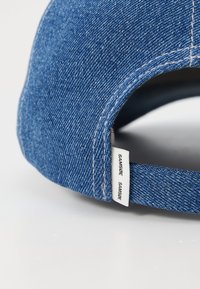 Gorra de denim con visera curvada, textura azul y etiqueta blanca cosida en la parte trasera con el nombre de la marca "Samsøe."