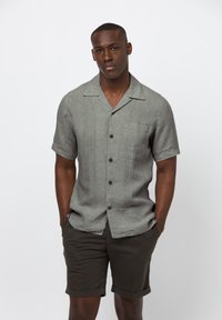 Korte mouwen, knoopsluiting shirt van lichtgrijze linnen met een borstzak en subtiele textuur, gecombineerd met donkerbruine shorts, handen in de zakken.