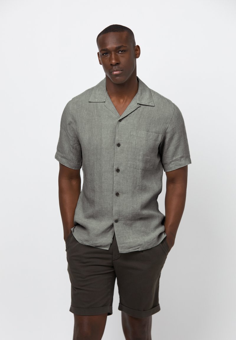 Korte mouwen, knoopsluiting shirt van lichtgrijze linnen met een borstzak en subtiele textuur, gecombineerd met donkerbruine shorts, handen in de zakken.