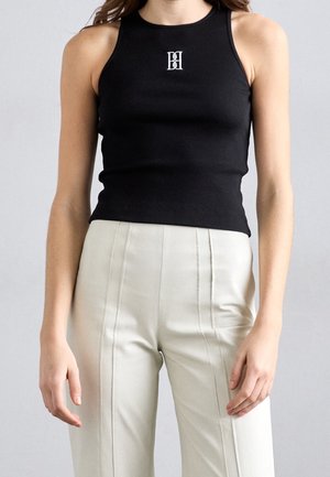 Femme portant un top noir sans manches côtelé avec logo blanc et un pantalon beige clair taille haute avec coutures visibles.