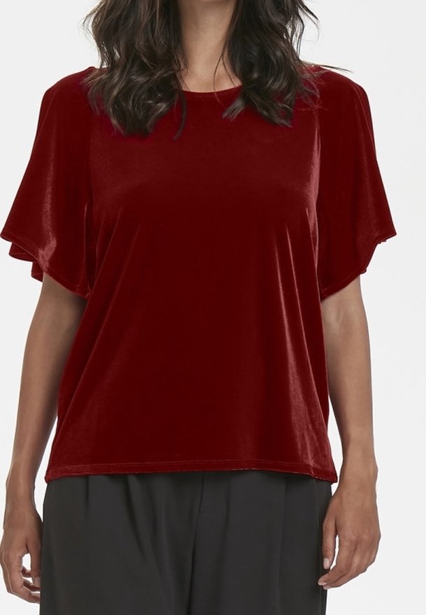 Femme portant un haut ample en velours rouge à manches courtes avec un col rond et un pantalon noir, sur un fond uni.