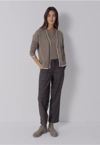 Lichtbruin vest met witte bies, grijze top, geruite broek in donkere tinten en beige enkellaarsjes. Model staat met handen in de zakken.