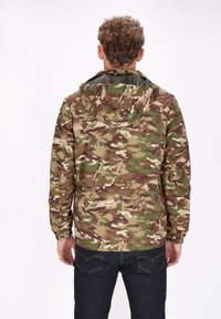 Giacca mimetica nelle tonalità di verde e beige, con cappuccio, polsini elasticizzati e tessuto leggero. Indossata con jeans in denim scuro.