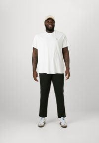 Camiseta blanca de manga corta con un pequeño logo verde, combinada con pantalones verdes oscuros y zapatillas blancas con detalles negros, de pie contra un fondo claro.