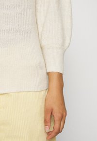 Osoba nosząca sweter z kremowego dzianiny z prążkowanymi mankietami oraz jasne żółte spodnie z korduroy, z ich zrelaksowaną ręką widoczną u boku.