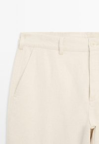 Pantalons en coton beige clair avec une coupe droite, des poches avant et une fermeture à bouton. Présente une texture subtile et des coutures soignées.