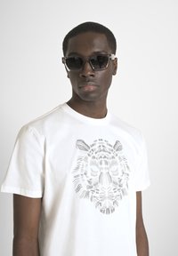 Camiseta blanca de algodón con un estampado gráfico de un tigre gris. El modelo lleva gafas de sol oscuras con un diseño de marco geométrico y lentes tintados.