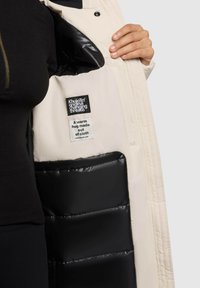 Persona che indossa una maglietta nera e tiene aperta una giacca beige chiara, rivelando un rivestimento imbottito nero e etichette cucite all'interno.