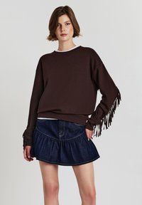 Felpa marrone con maniche frangiate, indossata sopra una camicia bianca. Abbinata a una gonna di denim blu con volant e una vita ben definita.
