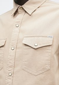 Bež srajca s gumbi in zloženo žepom, ki ima gumb na pritisek ter majhno oznako Jack & Jones na prsih.