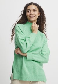Pulover verde dintr-un material tricotat moale, av�ând un guler înalt, manșete striate, slituri laterale și detalii texturate pe umeri.