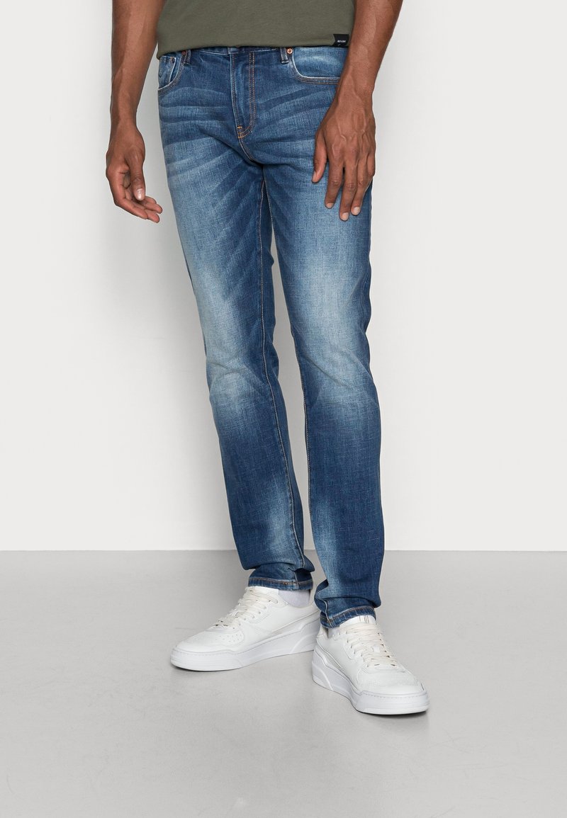 Vaqueros de mezclilla azul claro con un corte recto, lavado desgastado y detalles de costura naranja, combinados con zapatillas blancas.
