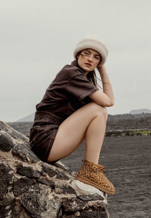 Botines de tobillo de estampado de leopardo en tono beige con un acabado suave, llevados con un mono oscuro, sentada en una roca contra un fondo gris.