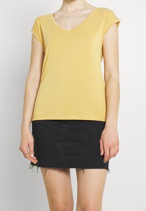 Mujer con una camiseta amarilla de manga corta y escote en V y una minifalda vaquera negra con dobladillo deshilachado sobre un fondo blanco liso.