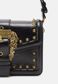 Svart läder crossbody-väska med guldfärgade nitar, blommiga dekorativa detaljer och en märkesplatta i metall på framfliken.