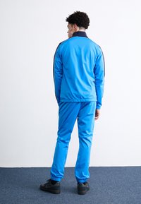 Μπλέ σύνολο με φερμουάρ και ταιριαστά παντελόνια, κατασκευασμένο από λείο ύφασμα, με ελαστικές μανσέτες και ενιαίο σχέδιο. Φοράει μαύρα παπούτσια.