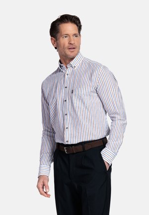 Man met bruin haar, gekleed in een gestreept overhemd met knopen, donkere broek en bruine riem, staand met één hand in de zak, kijkend naar links.