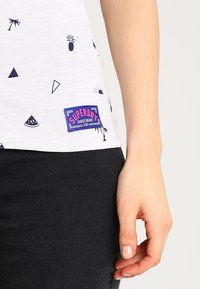 Camiseta blanca con estampados gráficos en azul marino de piñas, triángulos y sandías. Presenta un parche del logo "Superdry" en púrpura y rosa en el dobladillo.