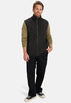 Schwarze und olivgrüne Fleece-Weste mit hohem Kragen, Reißverschluss und Fronttasche. Getragen mit schwarzen Hosen und Sneakers.