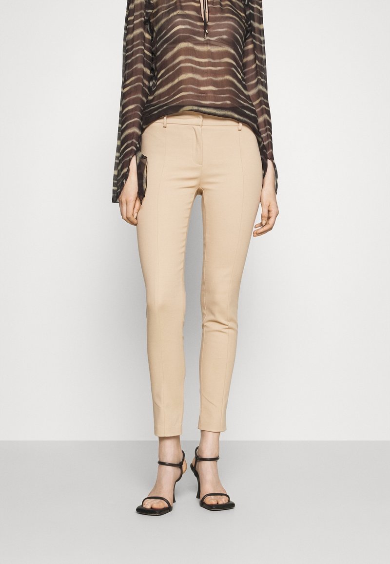 Femme portant un pantalon beige slim, des sandales à talons noires à brides et un haut marron rayé semi-transparent à manches longues sur un fond uni.