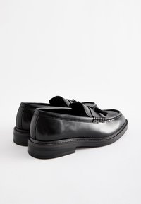 Mocassins en cuir noir présentant une finition lisse, une forme structurée, des coutures surélevées et des glands décoratifs à l'avant.