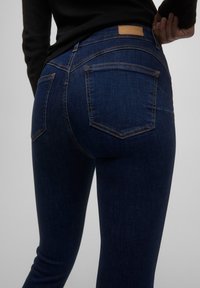 Jeans de denim azul oscuro con un diseño ajustado, que presentan bolsillos traseros y una etiqueta marrón en la cintura. Textura suave con costuras visibles.