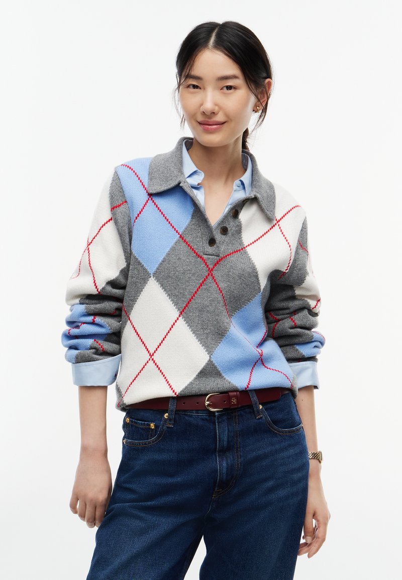 Femme portant une chemise bleue à col sous un pull à losanges gris, bleu et blanc avec des lignes rouges, un jean foncé et une ceinture marron, debout devant un fond blanc.