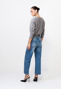 Femeie cu părul prins în coc, îmbrăcată cu un pulover gri supradimensionat, blugi albaștri cu talie înaltă, pantofi slingback negri cu toc înalt și cercei mari din argint.