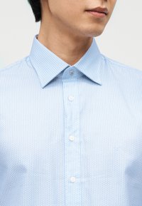 Άνδρας που φορά ένα γαλάζιο πουκάμισο με μικρό σχέδιο βέλους και γιακά, εμφανιζόμενος από τον λαιμό έως το μέσο του στήθους.