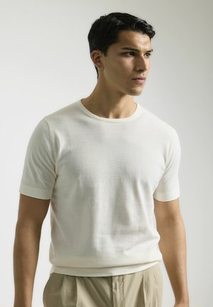 Suéter de punto corto de manga corta en color crema, con cuello redondo y puños acanalados, combinado con pantalones beige claros. Textura suave, diseño minimalista.
