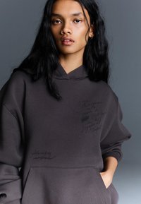 Hnedá mikina s kapucňou vyrobená z jemnej tkaniny, s prednou kapsou a potlačou v rukopisnom štýle na hrudi. Dizajn s kapucňou a rebrovanými manžetami.