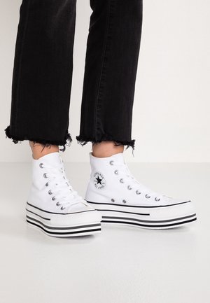 Osoba nosząca czarne spodnie z postrzępionym dołem i białe wysokie trampki Converse na platformie na białej podłodze i tle.