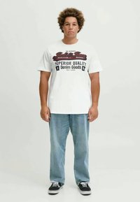Un t-shirt blanc avec un texte imprimé rouge et noir, associé à un jean bleu clair et des chaussures noires. Le t-shirt a une coupe décontractée et ample.