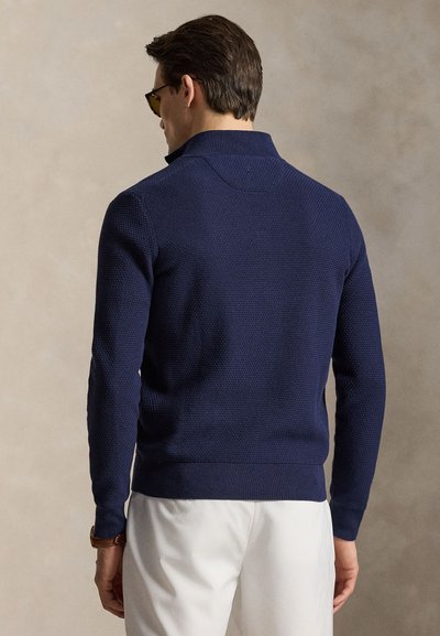 Férfi, aki tengerészkék, texturált hosszú ujjú pulóvert, fehér nadrágot, napszemüveget és barna karórát visel, háttal áll egy semleges háttér előtt.